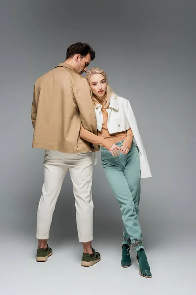 Full length of stylish model in pants and jacket leaning on man on grey — Stock Photo