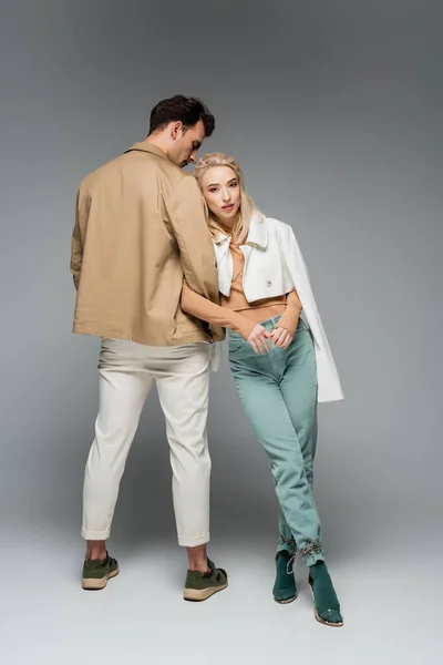 Longitud completa de modelo elegante en pantalones y chaqueta apoyada en el hombre en gris - foto de stock