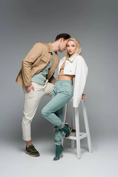 Longitud completa de los modelos de moda en pantalones y chaquetas posando cerca de la silla blanca en gris - foto de stock