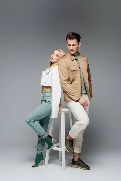 Longitud completa de elegante pareja en pantalones y chaquetas posando cerca de silla blanca en gris - foto de stock