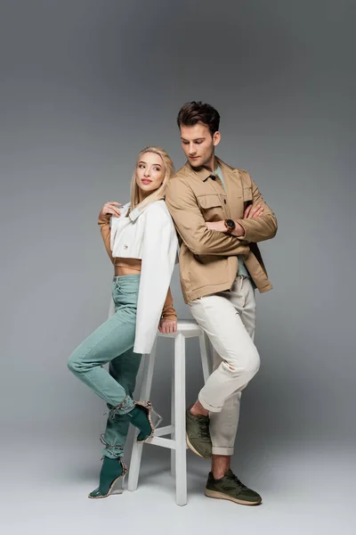 Comprimento total do jovem casal elegante em calças e jaquetas posando perto da cadeira branca no cinza — Fotografia de Stock