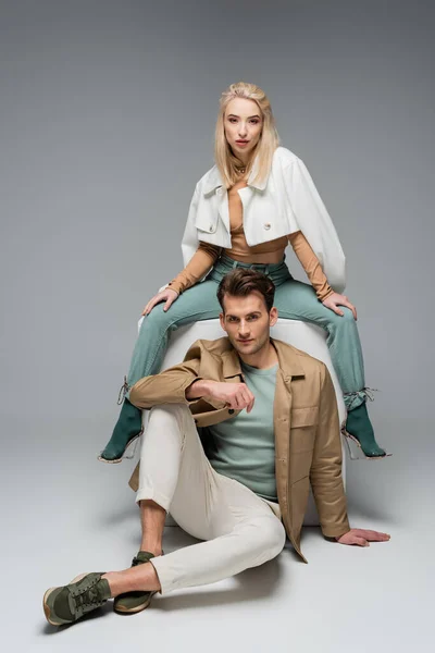 Full length of stylish models in pants and jackets posing near white cube on grey — Stock Photo