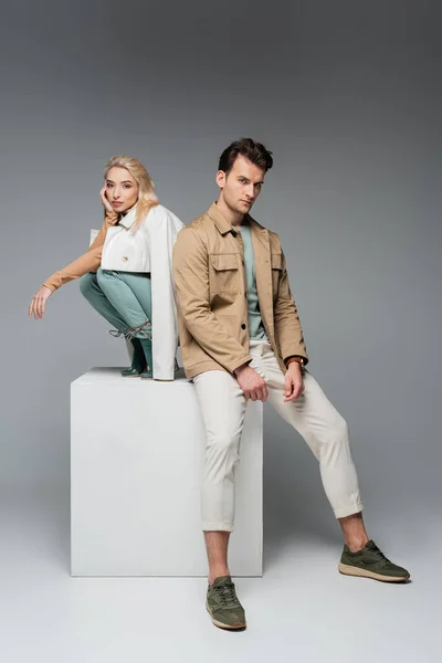 Full length of stylish models in pants and jackets posing while sitting on white cube on grey — Stock Photo