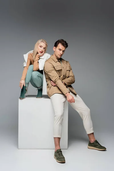 Full length of stylish man and woman jackets posing while sitting on white cube on grey — Stock Photo