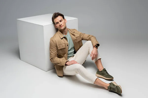 Full length of stylish man posing while leaning on white cube on grey — Stock Photo