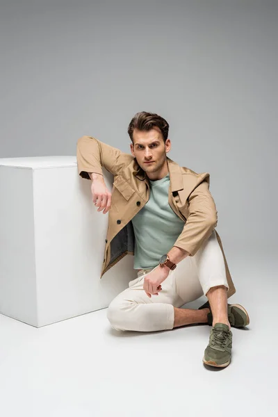 Full length of stylish man looking at camera while sitting near white cube on grey — Stock Photo