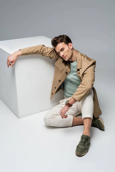 Full length of trendy man posing while leaning on white cube on grey — Stock Photo