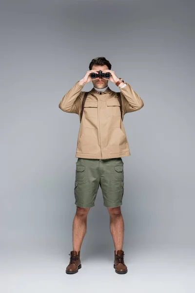 Tutta la lunghezza del viaggiatore alla moda in pantaloncini corti e giacca guardando attraverso binocoli su grigio — Foto stock