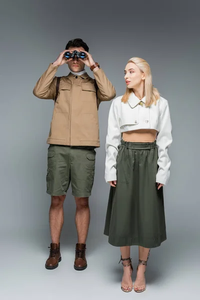 Comprimento total do viajante na moda olhando através de binóculos perto de mulher loira em cinza — Fotografia de Stock
