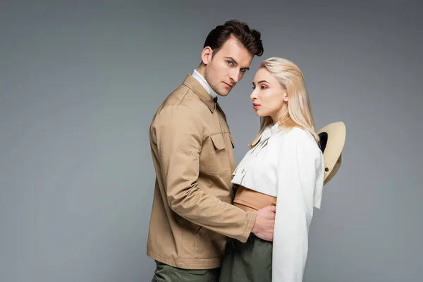 Hombre con estilo en la chaqueta abrazando mujer joven rubia aislada en gris - foto de stock