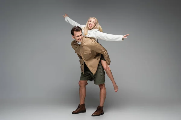 Full length of happy man piggybacking cheerful blonde woman on grey — Stock Photo