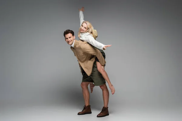 Full length of cheerful man piggybacking cheerful blonde woman on grey — Stock Photo