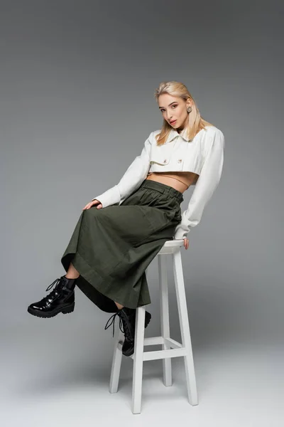 Full length of blonde model in cropped jacket and skirt sitting on white chair on grey — Stock Photo