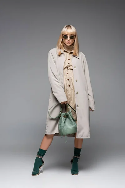 Longitud completa de la mujer joven de moda en gafas de sol y gabardina posando con bolsa verde en gris - foto de stock