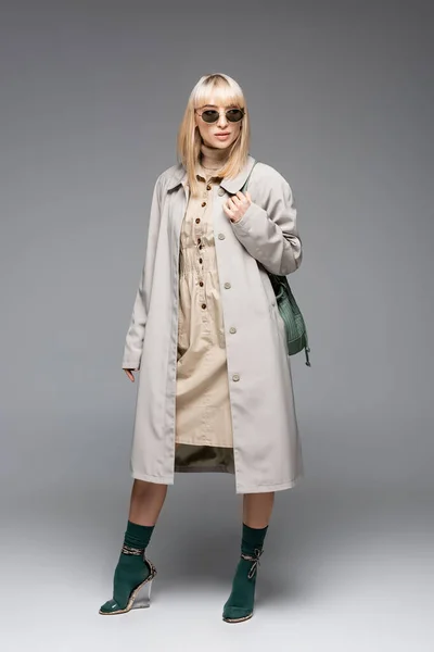 Pleine longueur de femme à la mode en lunettes de soleil et trench coat posant avec sac vert sur gris — Photo de stock