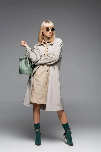 Pleine longueur de femme à la mode en lunettes de soleil et trench coat posant avec sac vert tout en se tenant debout sur le gris — Photo de stock