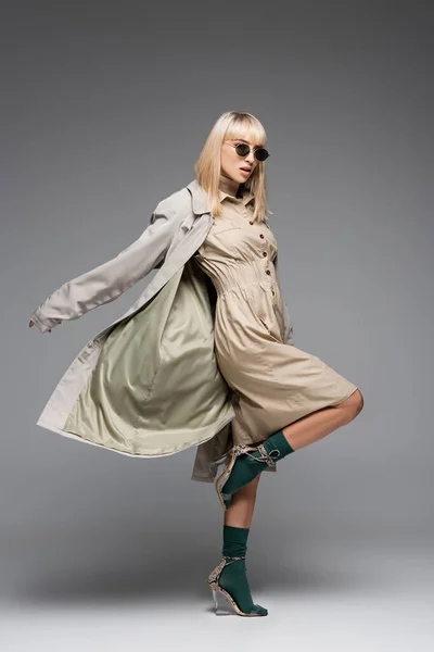 Pleine longueur de femme à la mode en lunettes de soleil et trench coat posant sur gris — Photo de stock