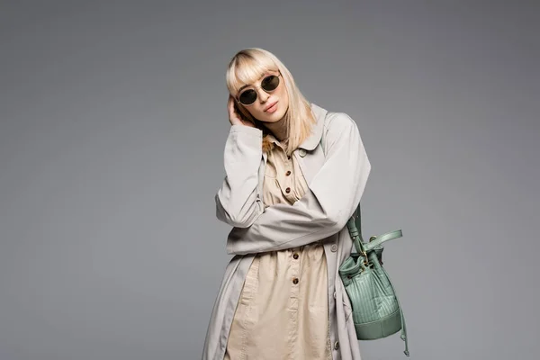 Donna alla moda in occhiali da sole e trench in posa con borsa verde mentre in piedi isolato su grigio — Foto stock