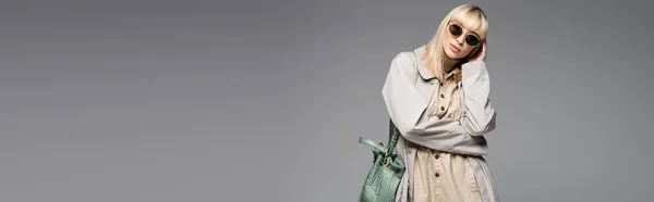 Модная женщина в солнцезащитных очках и плаще позирует с зеленой сумкой, стоя изолированно на сером, баннер — стоковое фото