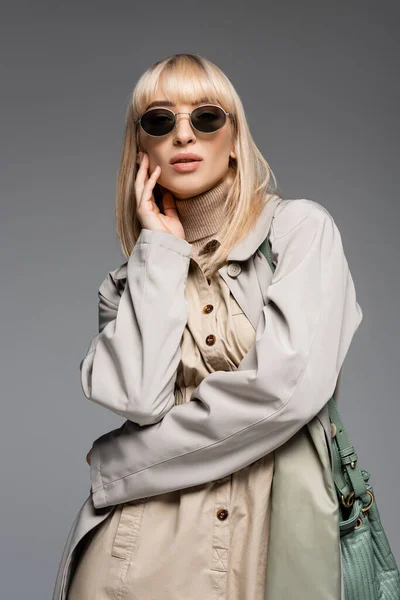 Joven mujer de moda en gafas de sol y gabardina posando aislado en gris - foto de stock