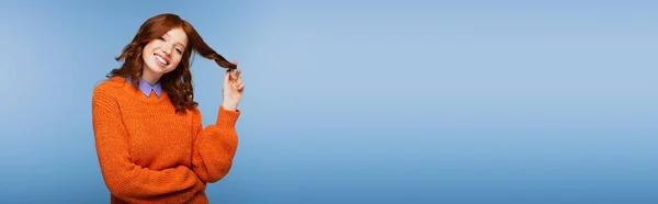 Mulher ruiva feliz em suéter laranja curling cabelo no dedo isolado no azul, banner — Fotografia de Stock