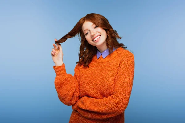 Happy redhead woman in orange sweater curling hair on finger isolated on blue — Stock Photo