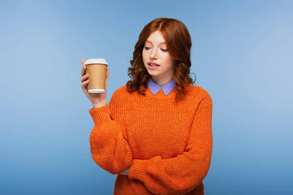 Pelirroja en suéter naranja sosteniendo café para llevar mientras muerde los labios aislados en azul - foto de stock