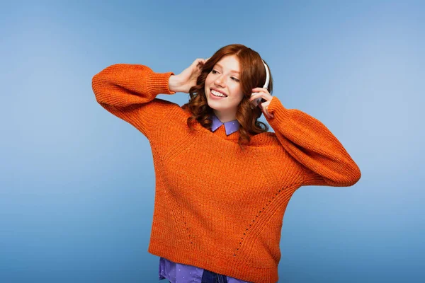 Glücklich rothaarige Frau hört Musik in drahtlosen Kopfhörern isoliert auf blau — Stockfoto