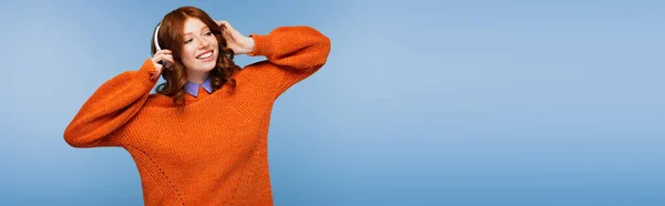 Mujer pelirroja feliz escuchando música en auriculares inalámbricos aislados en azul, bandera - foto de stock