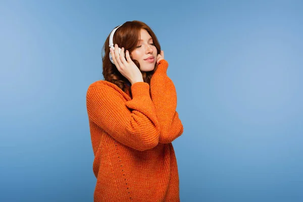 Heureux rousse femme avec les yeux fermés écouter de la musique dans les écouteurs sans fil isolé sur bleu — Photo de stock