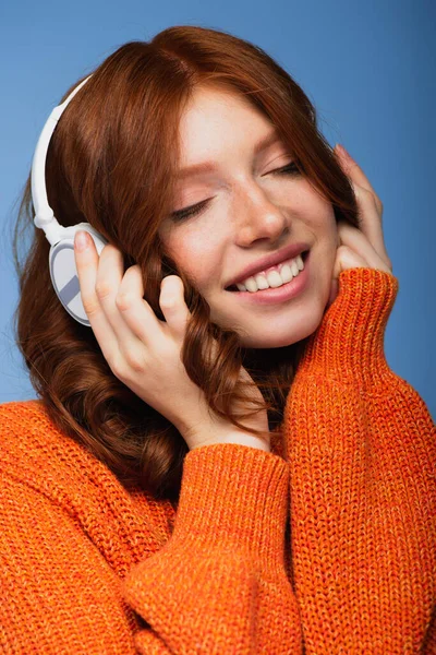 Happy redhead woman with closed eyes listening music in wireless headphones isolated on blue — Stock Photo