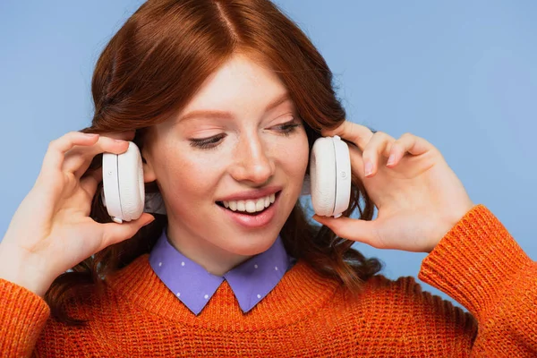 Mujer pelirroja feliz sosteniendo auriculares inalámbricos aislados en azul - foto de stock