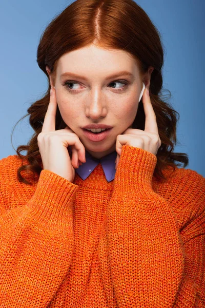 Redhead woman listening music while touching wireless earphones isolated on blue — Stock Photo