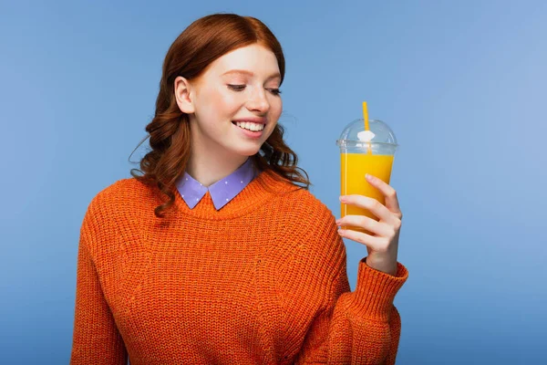 Glücklich rothaarige Frau im Pullover hält frischen Orangensaft in Plastikbecher isoliert auf blau — Stockfoto