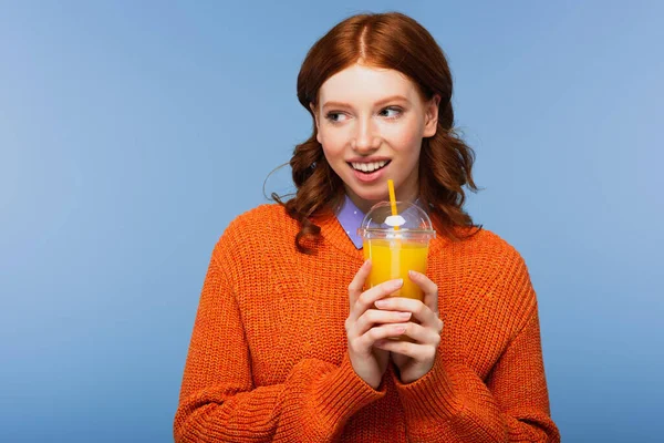 Sonriente pelirroja en suéter sosteniendo jugo de naranja fresco en taza de plástico aislado en azul - foto de stock