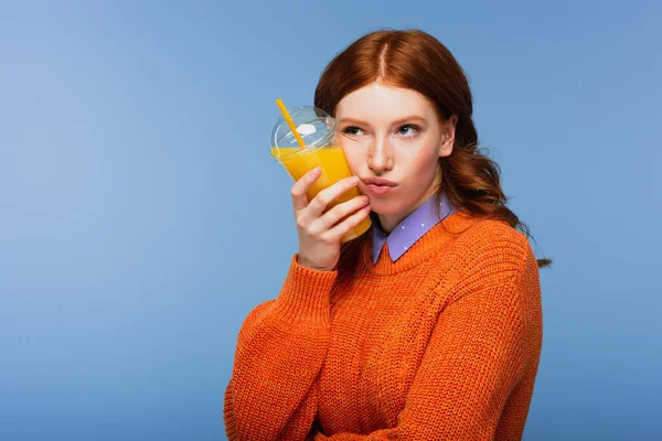 Bored redhead woman in sweater holding fresh orange juice in plastic cup isolated on blue — Stock Photo