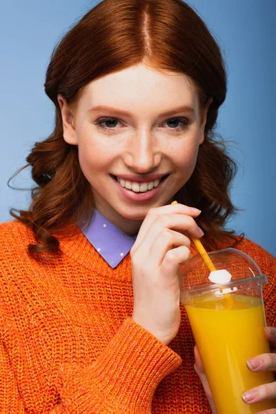Feliz mujer joven en suéter sosteniendo jugo de naranja fresco en taza de plástico aislado en azul - foto de stock