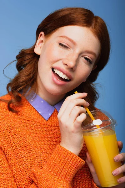 Feliz pelirroja mujer en suéter celebración de jugo de naranja fresca en taza de plástico mientras guiño ojo aislado en azul - foto de stock