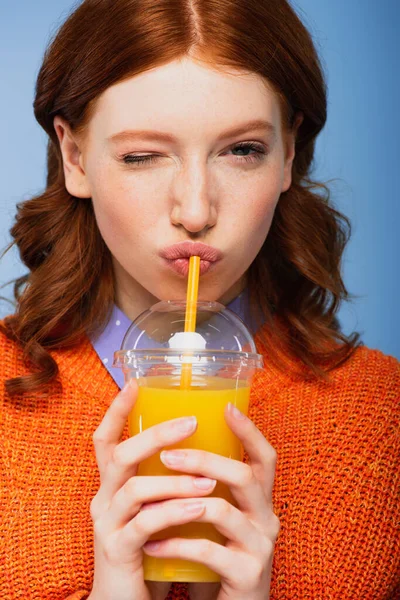 Redhead woman in sweater drinking fresh orange juice isolated on blue — Stock Photo