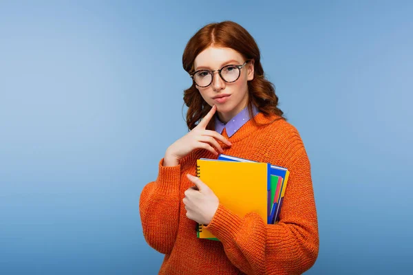 Estudiante pensativo en gafas y suéter naranja con cuadernos aislados en azul — Stock Photo