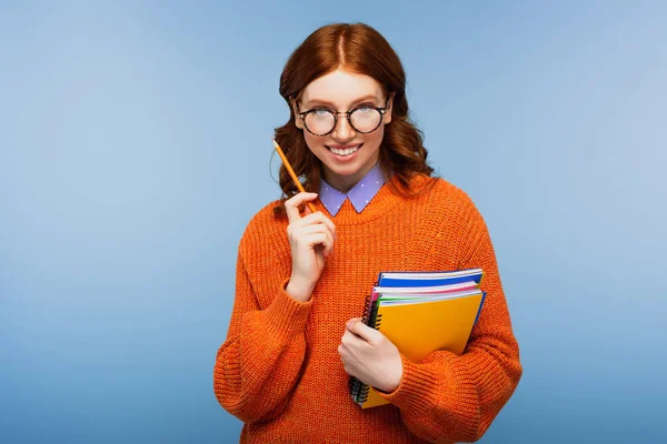 Heureux jeune étudiant en lunettes et pull orange tenant des carnets et crayon isolé sur bleu — Photo de stock