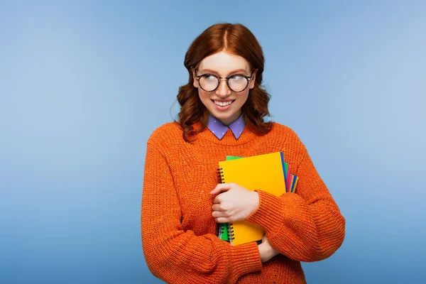 Estudiante pelirroja sonriente en gafas y suéter naranja sosteniendo cuadernos y lápiz aislado en azul - foto de stock