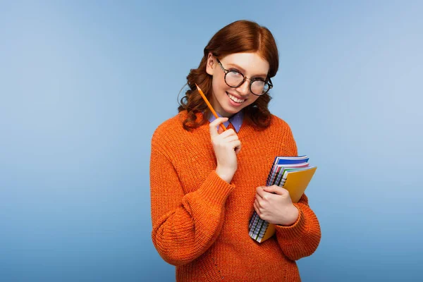 Весела руда студентка в окулярах і помаранчевий светр тримає блокноти і олівець ізольовані на синьому — стокове фото