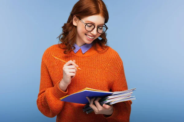 Estudiante feliz en gafas y suéter sosteniendo lápiz y cuadernos aislados en azul - foto de stock