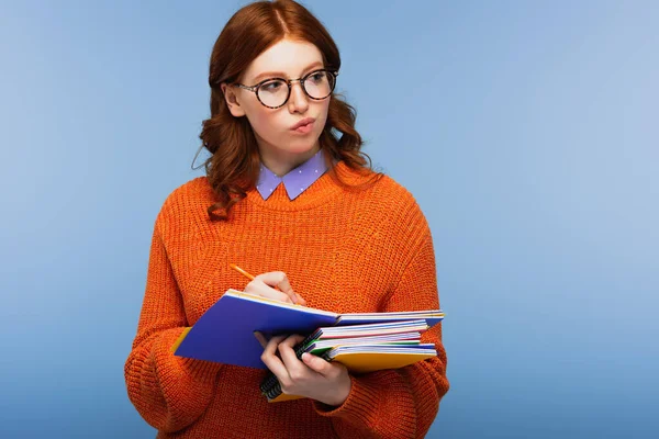 Estudiante reflexivo en gafas y suéter sosteniendo lápiz y cuadernos aislados en azul - foto de stock