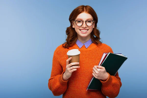 Alegre joven estudiante en gafas y suéter naranja sosteniendo cuadernos y taza de papel aislado en azul — Stock Photo