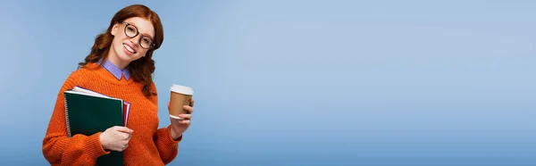 Felice studente rossa in occhiali e maglione arancione che tiene quaderni e bicchieri di carta isolati su blu, banner — Foto stock