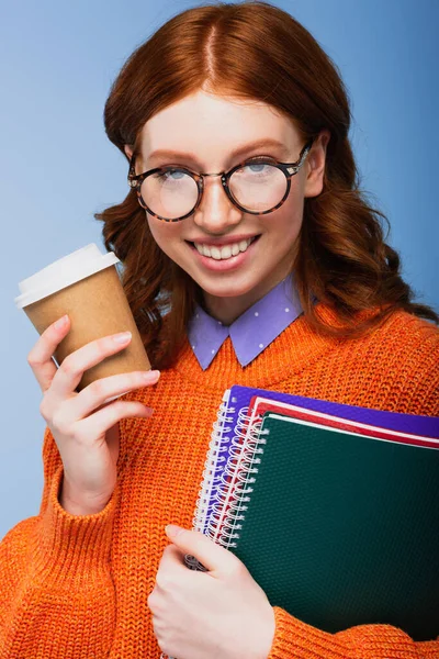 Estudante feliz em óculos e suéter laranja segurando cadernos e copo de papel isolado em azul — Fotografia de Stock