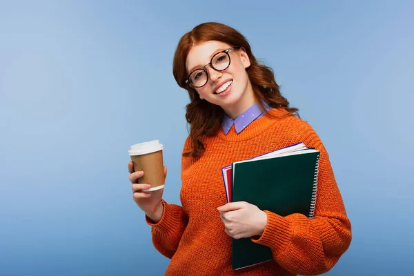 Estudiante pelirroja feliz en gafas y suéter naranja sosteniendo cuadernos y taza de papel aislado en azul - foto de stock