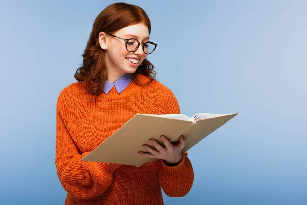 Estudante ruiva feliz em óculos e camisola laranja leitura livro isolado em azul — Fotografia de Stock